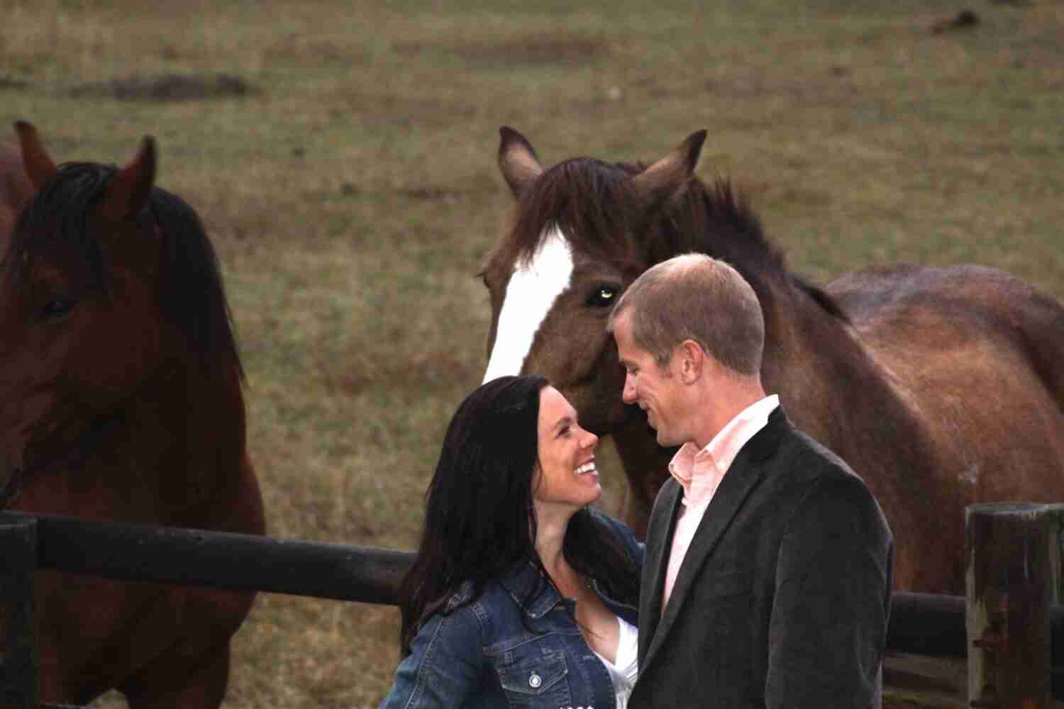 a couple smiling at each other with horses in a field